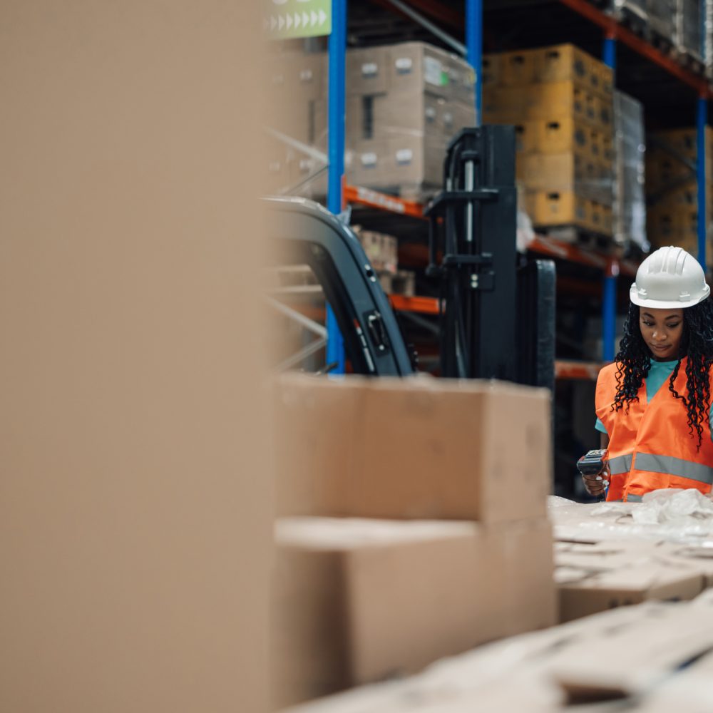 Warehouse worker is scanning barcodes on packages while working in a busy distribution center. She is wearing a safety vest and helmet