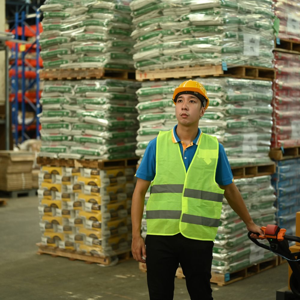 Male warehouse worker in hardhats and reflective jackets pulling a pallet truck walking through rows of storage racks with merchandise.