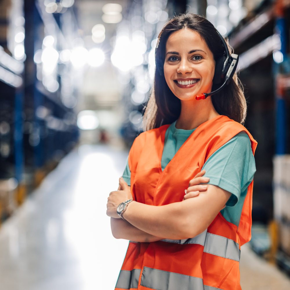 A confident female warehouse worker stands with arms crossed, wearing a headset in a brightly lit industrial space, symbolizing confidence and readiness in logistics work.